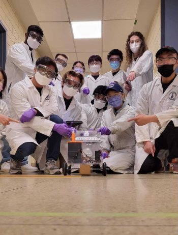 A group of students in white lab coats pose around a model-sized car cars powered by chemical energy sources.
