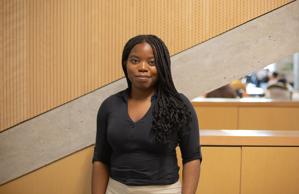 A woman with long black braids and black shirt stands in front of a panelled wall and railing.