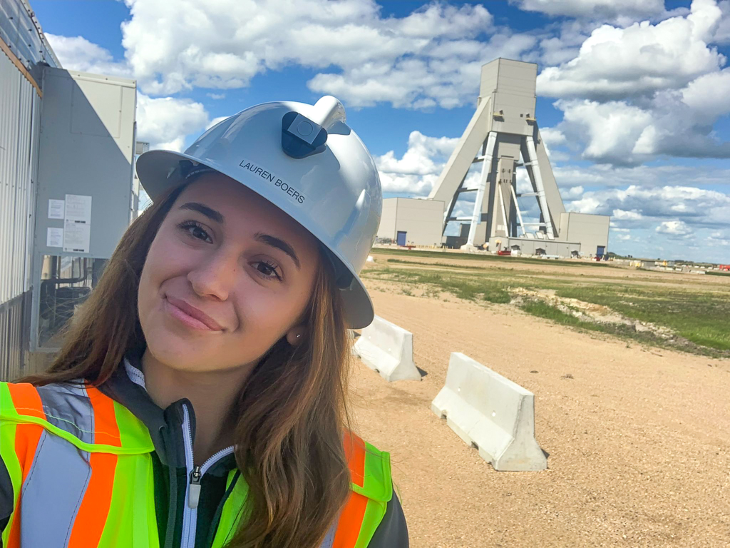 A smiling woman is seen from the shoulders up. She is wearing a white hard hat and fluorescent safety vest. Behind her is a mining site.