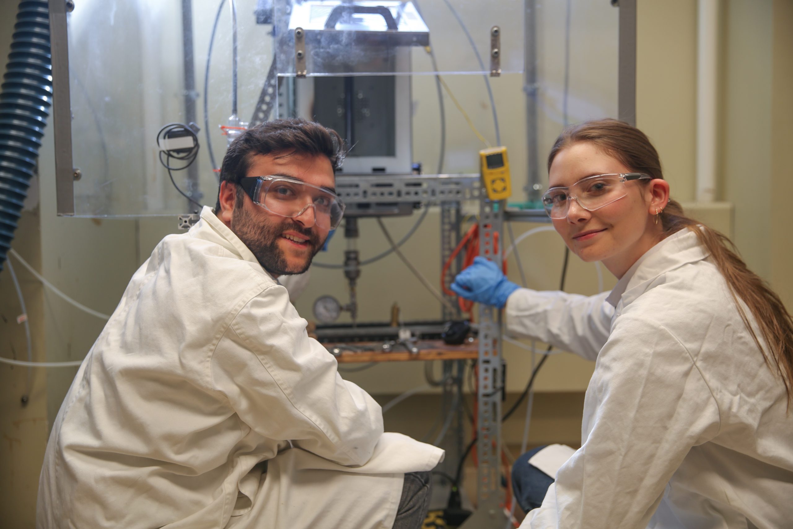 Two researchers wearing protective lab coats, gloves and googles look back while working in a laboratory.