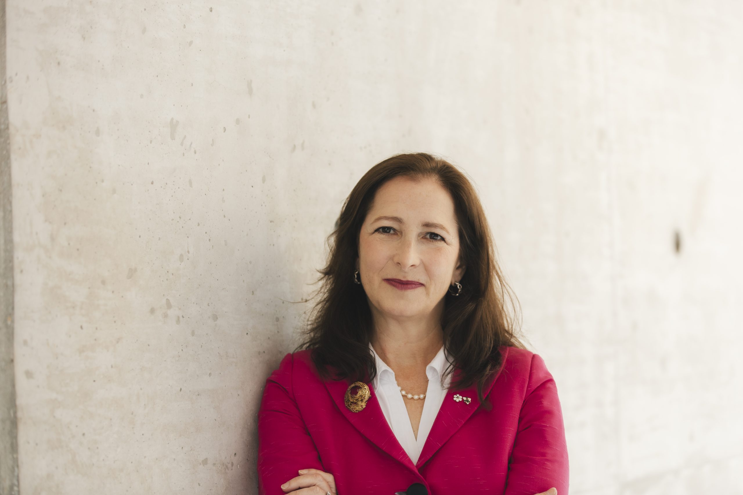 Woman with brown hair and red blazer stands in front of a wall.