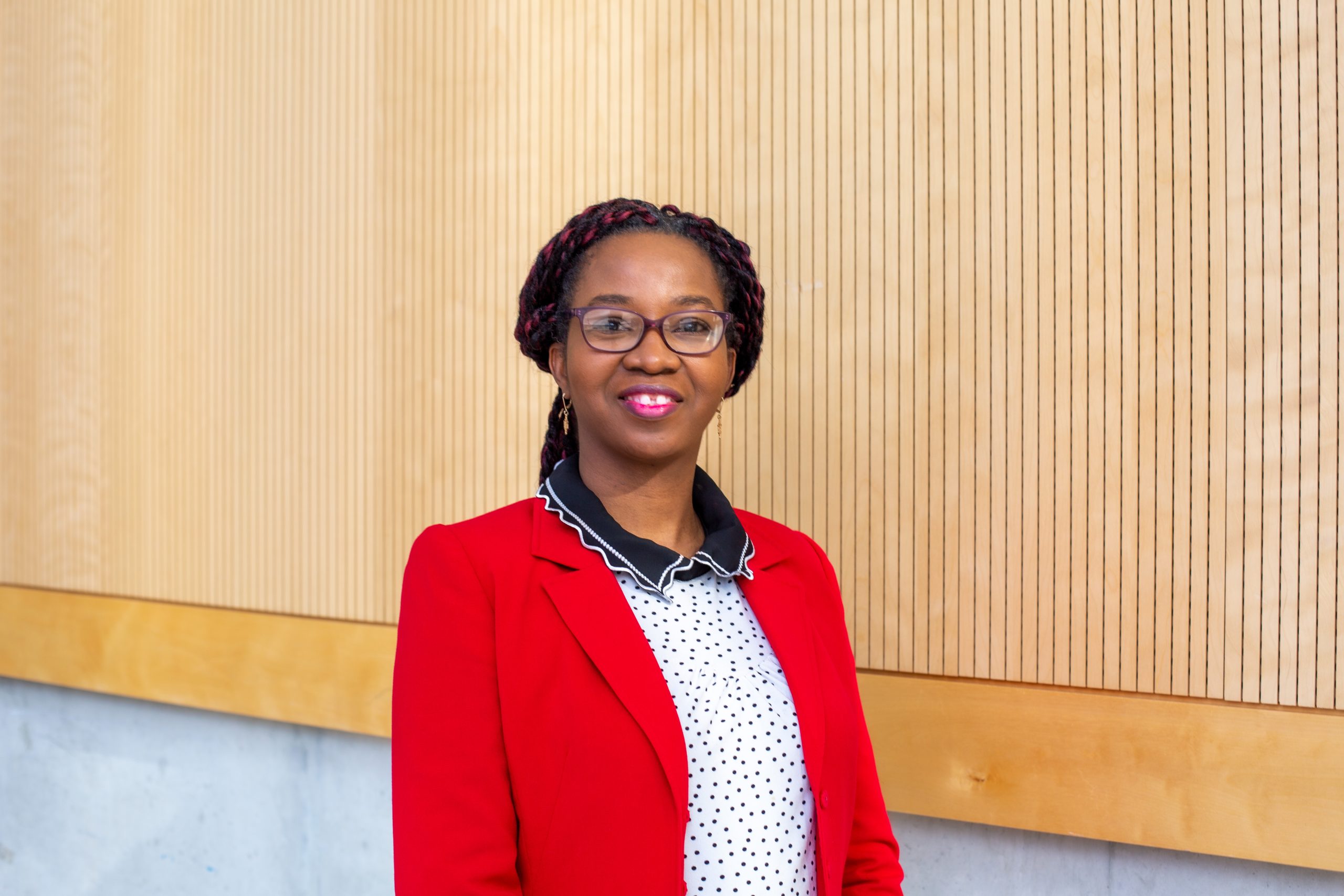 A woman in a red blazer and glasses stands in front of wood-panelled wall.