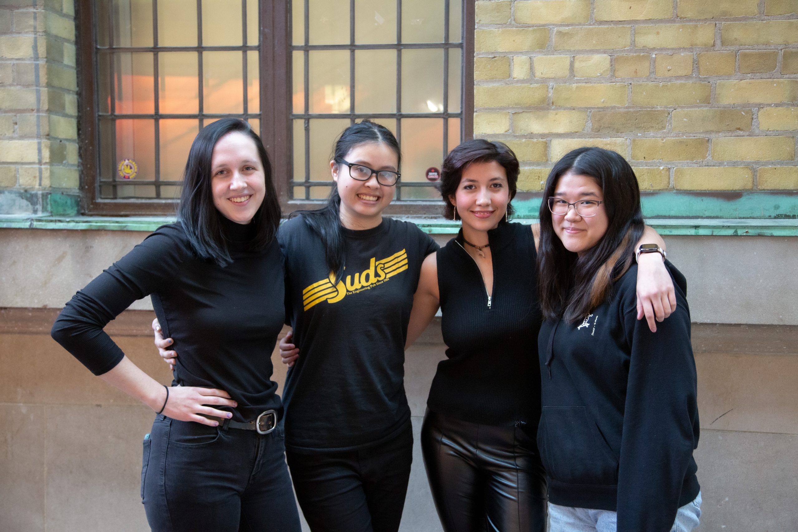 Four women stand together in front a wall with bricks and a window.