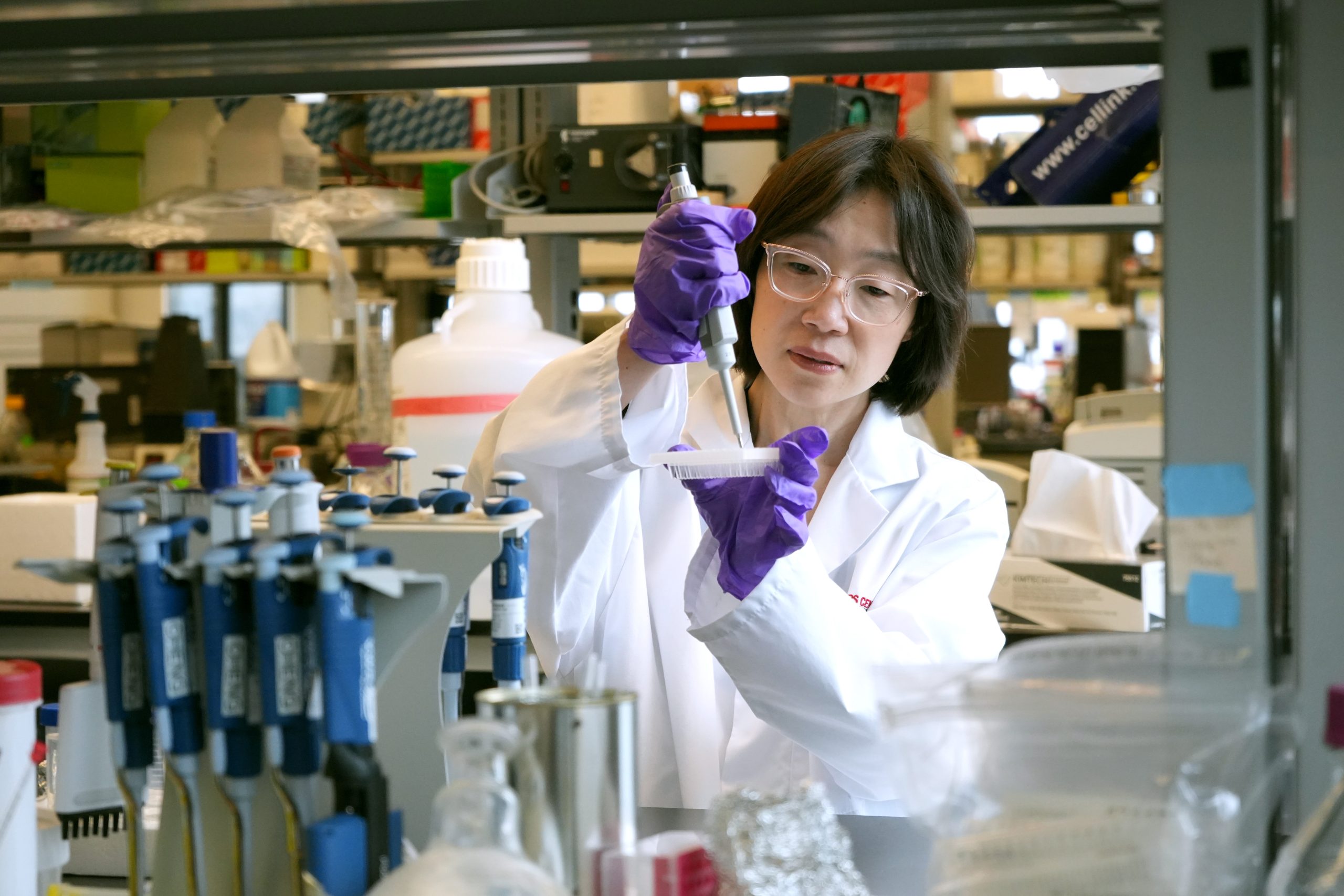 Hai-Ling Cheng is wearing a lab coat and pipetting a solution into a dish. Cheng appears to be in a lab setting. Pipettes and boxes are visible.