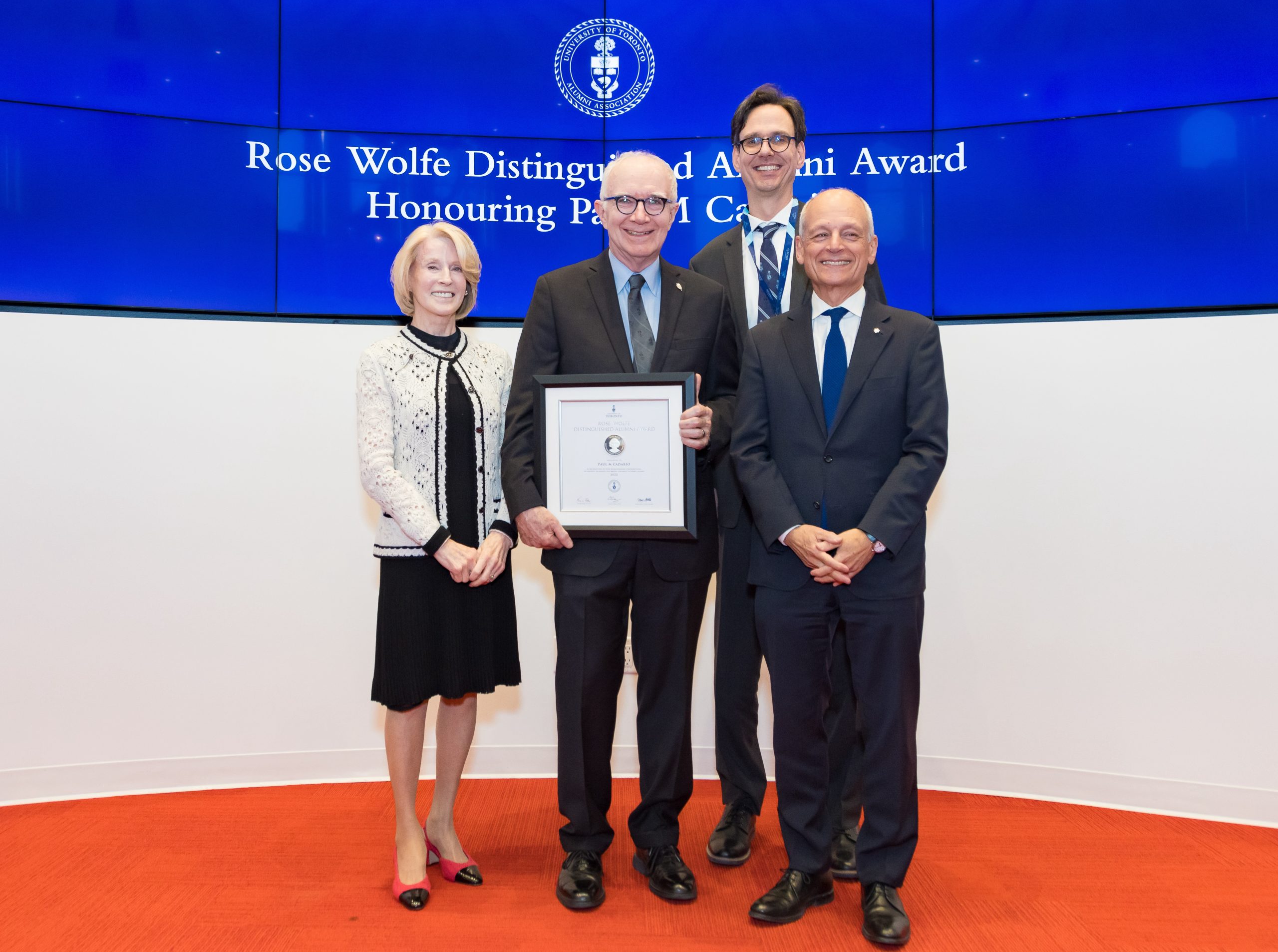 Paul Cadario stands in the centre holding his award certificate in front of him. To Cadario's left stands President Meric Gertler. To Cadario's right stands Chancellor Rose Patten. UTAA President Corwin Cambray stands at the back. A sign behind them has the U of T crest and reads: Rose Wolfe Distinguished Alumni Award. Honouring Paul Cadario.