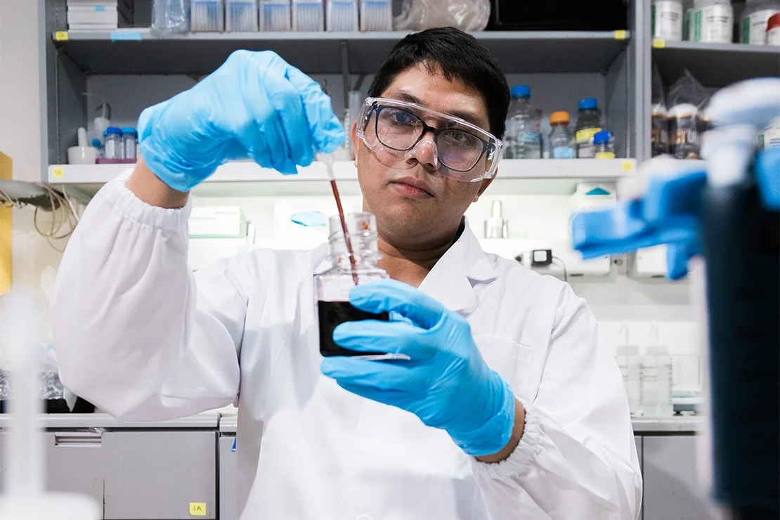 Pratish Gawand pipettes a coloured liquid in a beaker. Gaward, who appears to be in a lab setting, is dressed in a white lab coat.