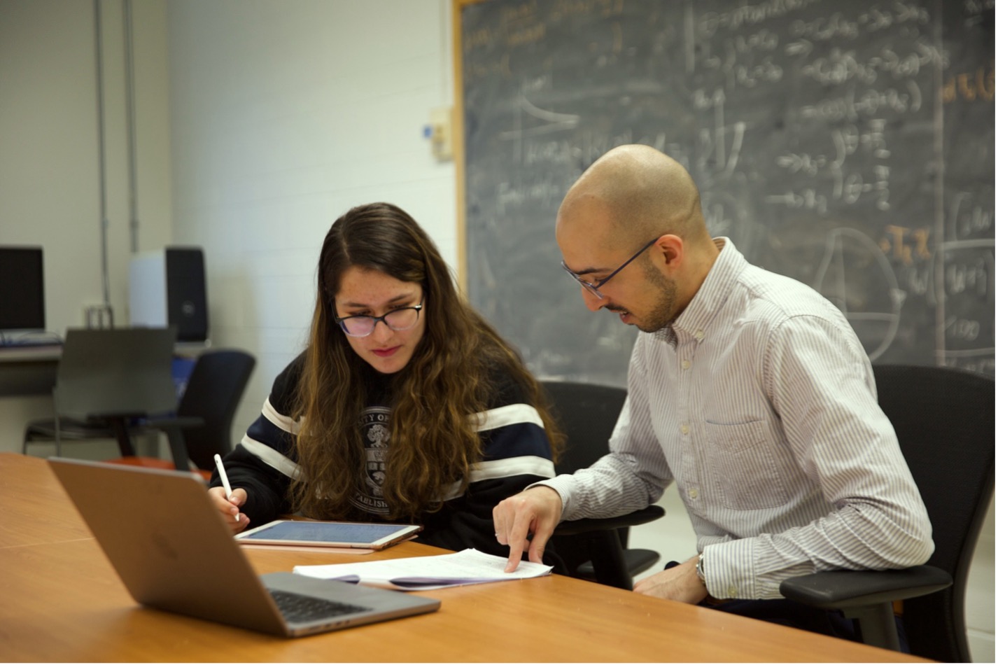 From left to right: PhD student Niloufar Yousefi is seated next to Professor John Simpson-Porco in a classroom setting. They are both looking at material on their shared workspace. There is black chalk board in the background. (photo by Matthew Tierney)