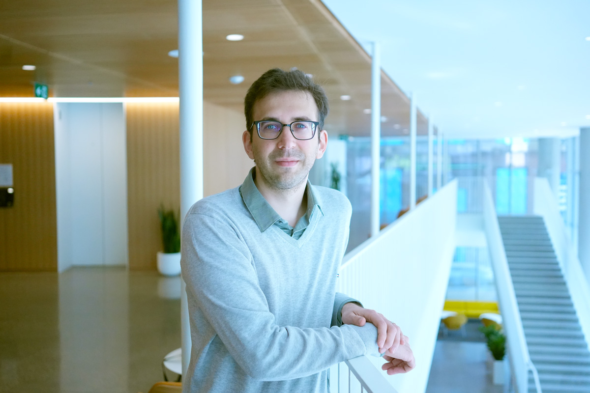 Professor Papernot stands in the hallway inside a building leaning on a railing that looks to the lower level. In the background is a stairwell to the right and an elevator door on the left.