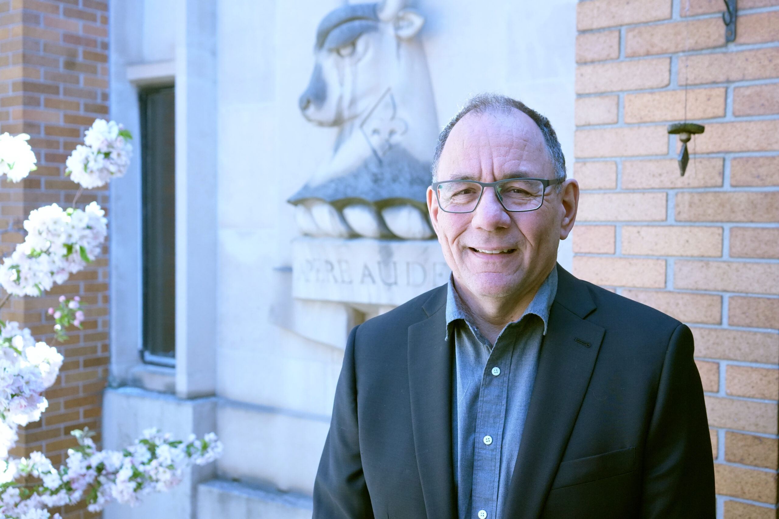 A close portrait of Jonathan Rose wearing a dark blazer over a blue dress shirt. He is standing in front a building and next to blossoms.