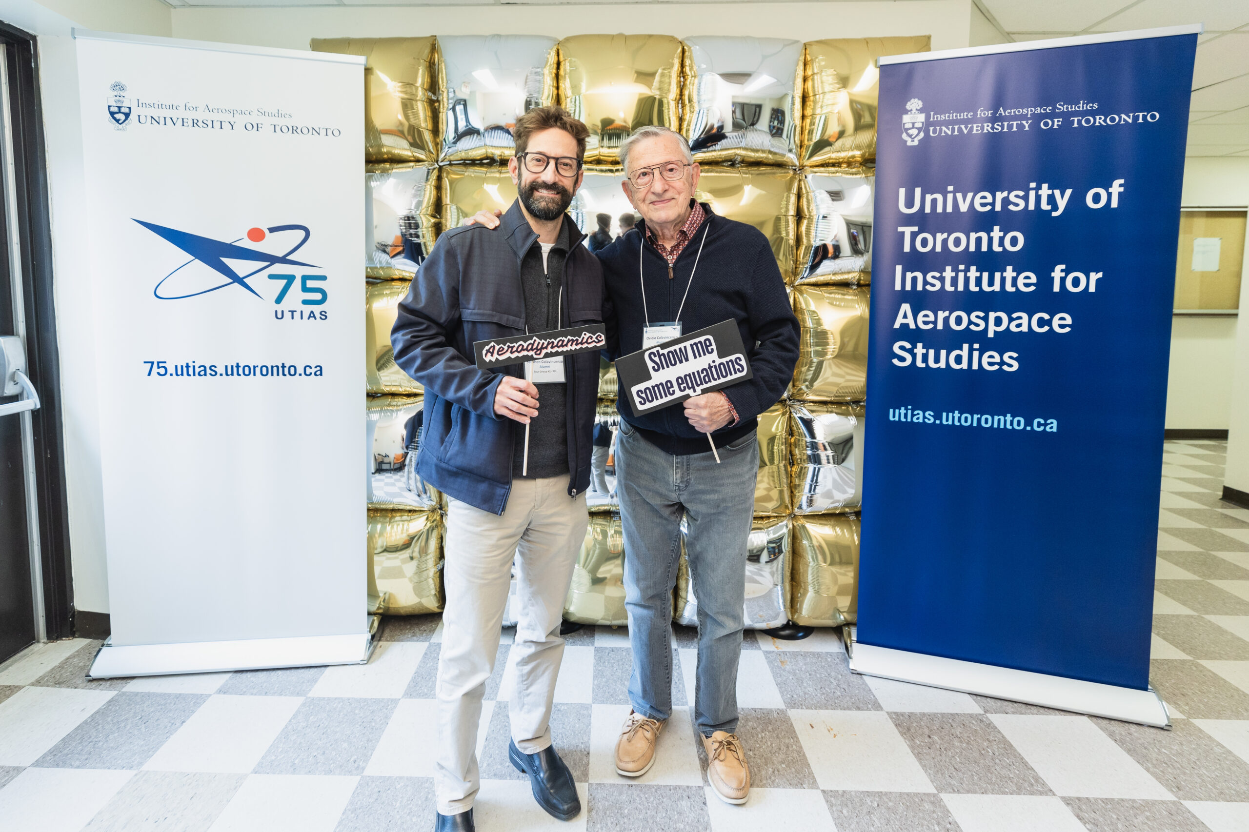 Two individuals pose between two banners, one has a logo with the words 75 UTIAS, the other reads University of Toronto Institute for Aerospace Studies. Behind them is a background of square balloons in gold and silver.