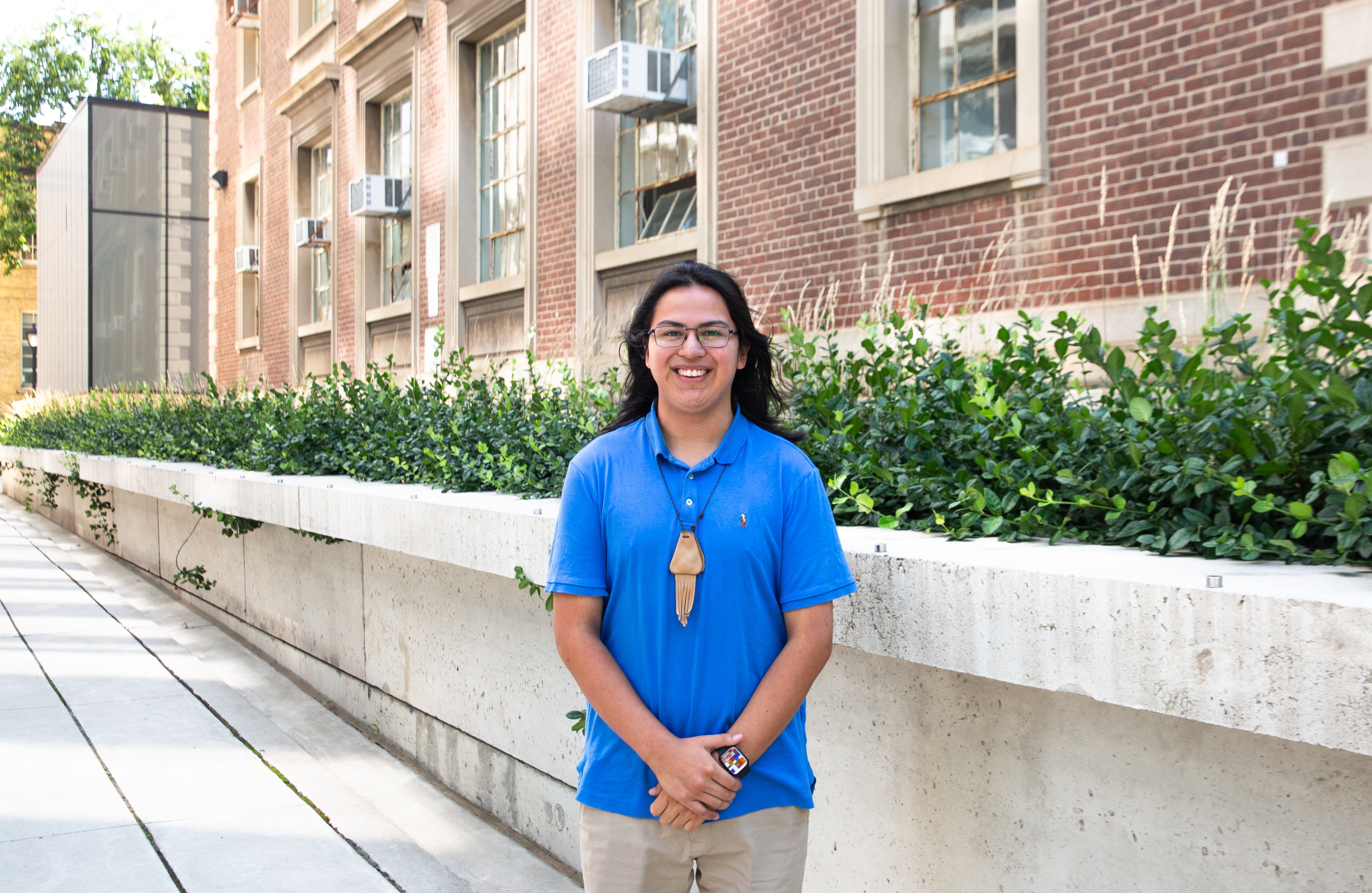 Connor Isaac stands holding his arms in front of him. He's wearing a light blue golf shirt and khaki pants with a pendant around his neck. Behind him is a building with a row of plants in front of it.