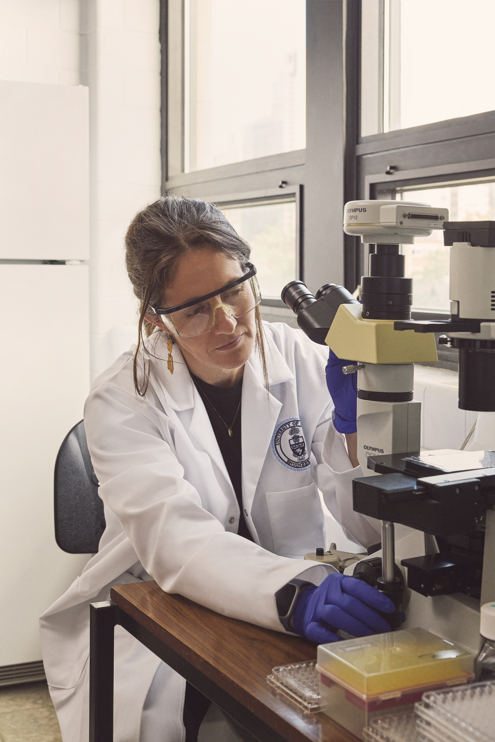 Professor McGuigan wears a white lab coat, safety goals and blue gloves while working in her lab. 