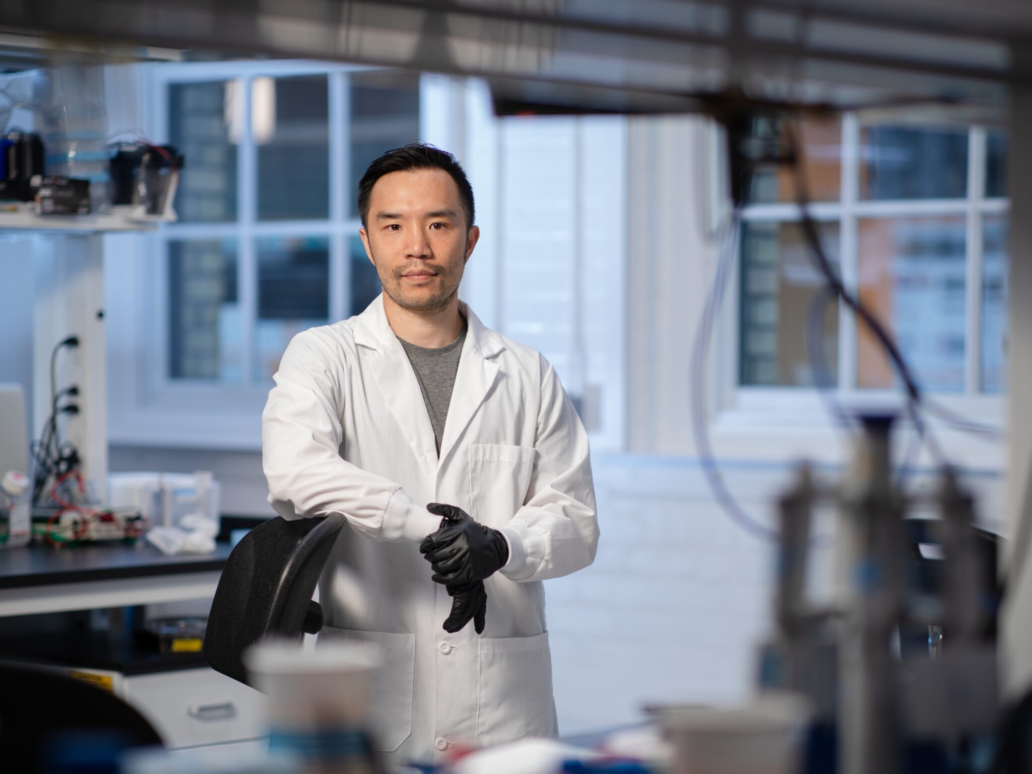 Professor Leo Chou is in a lab coat and gloves, leaning on a chair in his lab. He is looking at the camera.