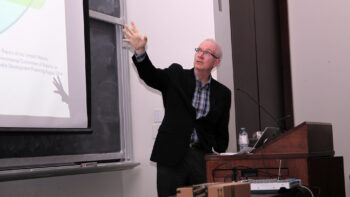 Paul speaks and points at a screen, while standing in front of a podium.