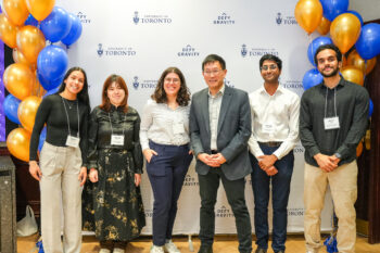 A group of 5 students and Dean Chris Yip pose for a photo in front of a U of T banner flanked by balloons.