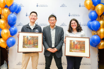 Two students holding framed pictures pose with Dean Chris Yip in front of a U of T banner flanked by balloons.