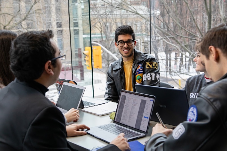 Students sit at a table with laptop computers. One student is in focus; he wears a leather jacket with a yellow sweater underneath. Floor-to-ceiling windows behind them give a view of trees and a traffic light.