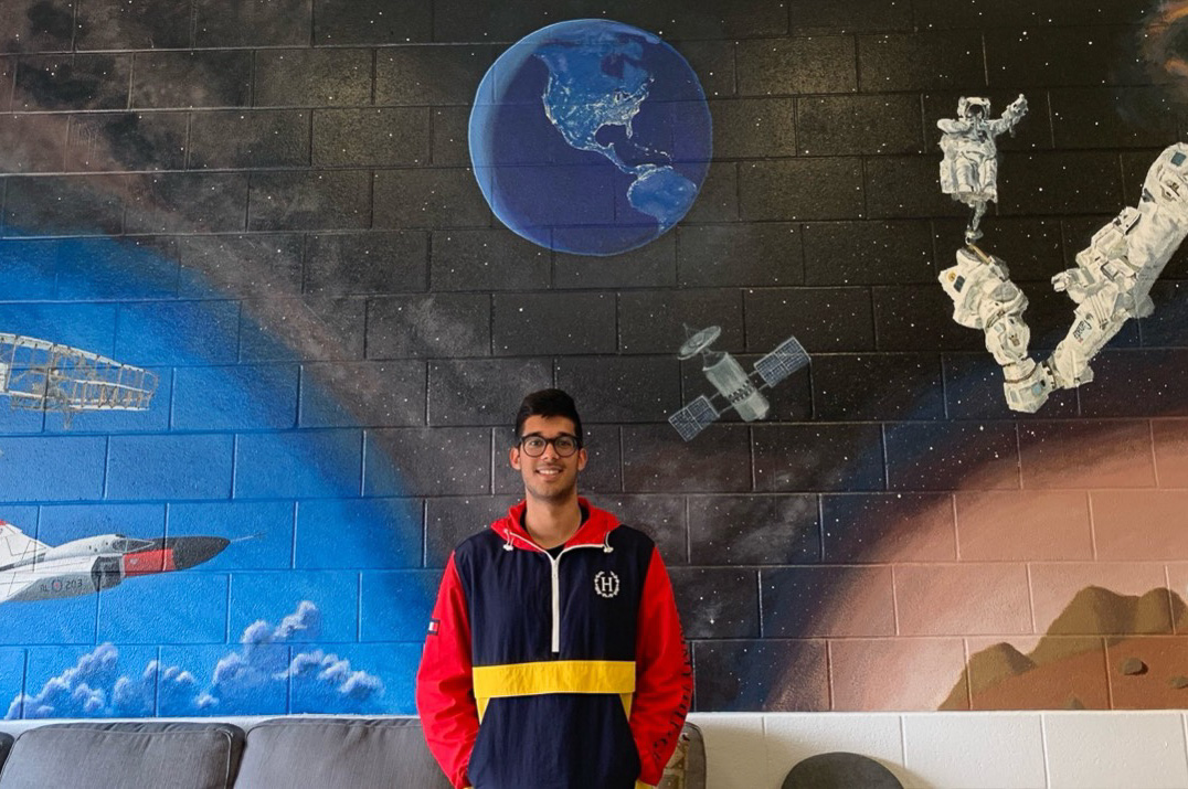 A man in a multi-coloured half-zip sweater stands in front of a large mural painting depicting scenes from space research.