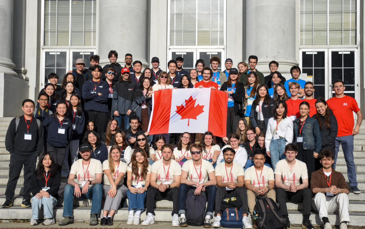 A large group of students pose on steps with a Canadian flag held in the middle of the group