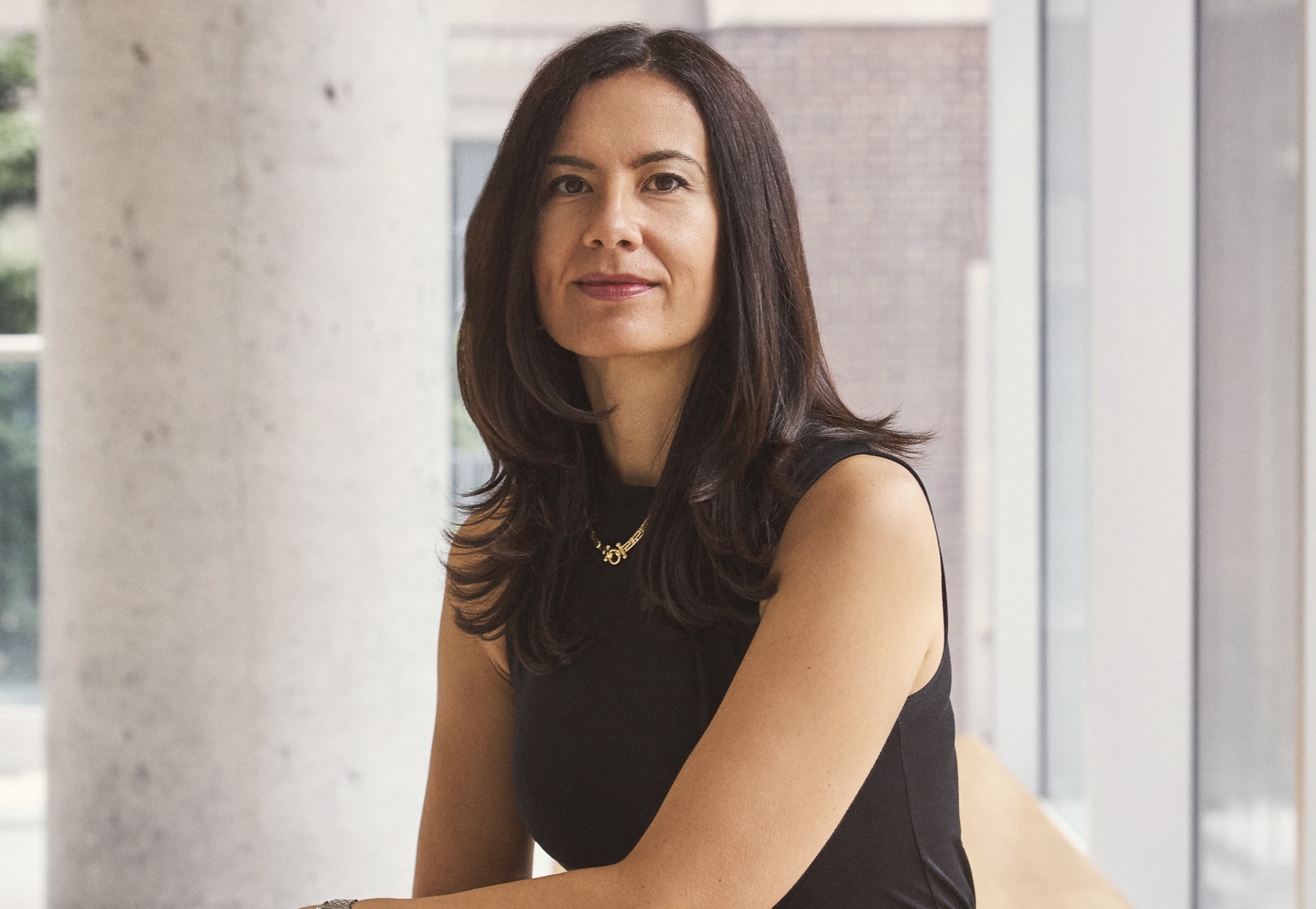 Milica sits on a bench, smiles at the camera. A concrete pillar, walls and glass panelling are in the background.