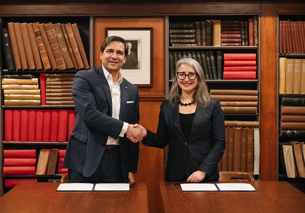 Kazi is on the left, dressed in a black suit, shaking hands with Cowen on the right, also in a black suit. Both stand in front of a bookshelf, with a table in front of them and the agreements on the table.