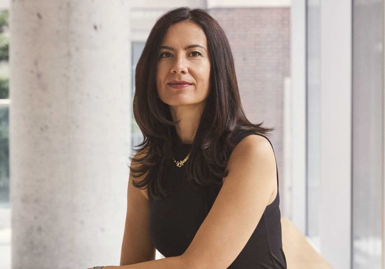 Milica sits on a bench, smiles at the camera. A concrete pillar, walls and glass panelling are in the background.