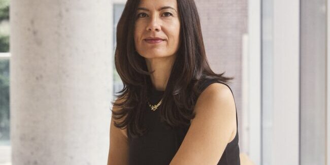 Milica sits on a bench, smiles at the camera. A concrete pillar, walls and glass panelling are in the background.