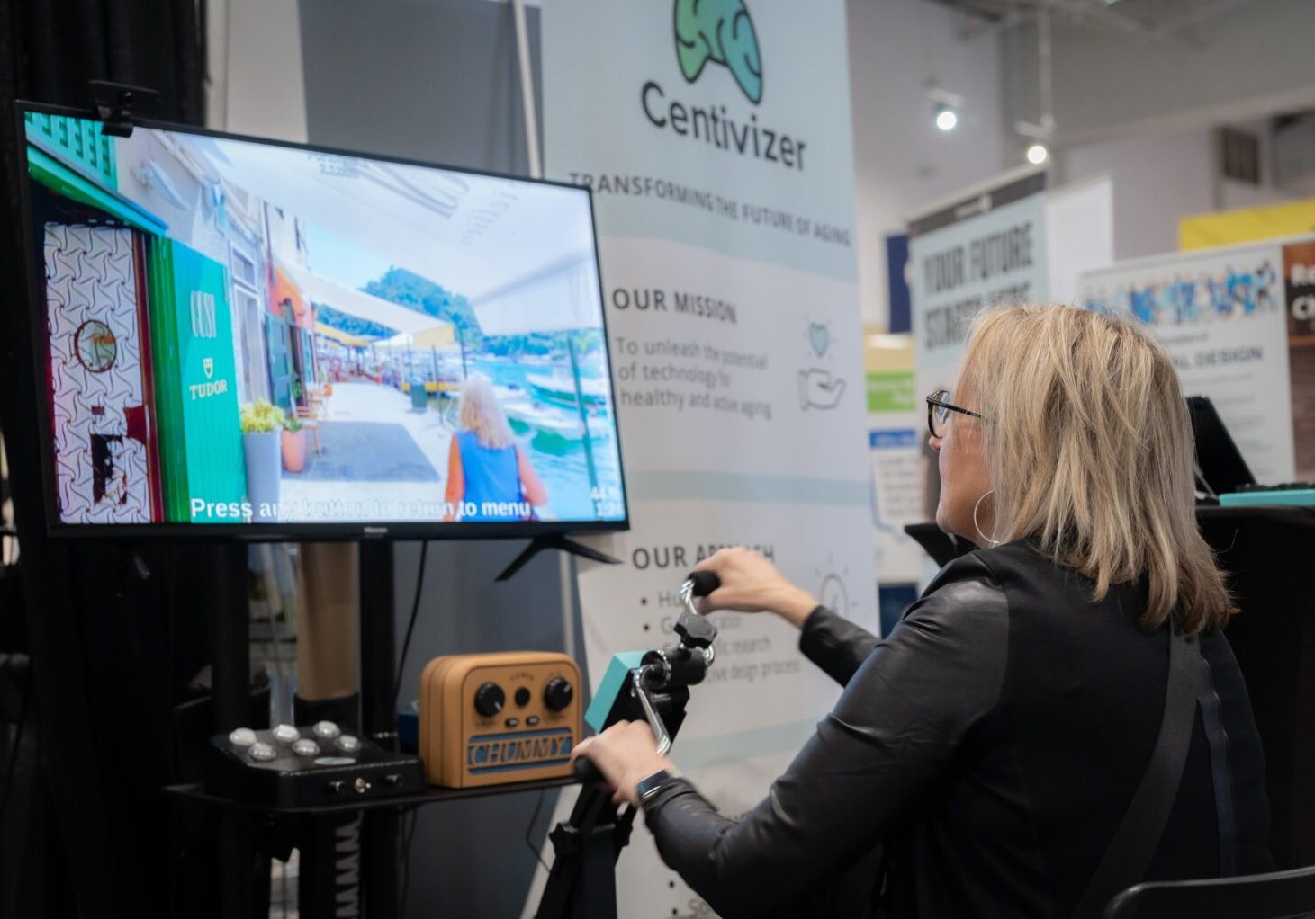 A woman uses a seated exercise device with a screen featuring a woman walking on the street.
