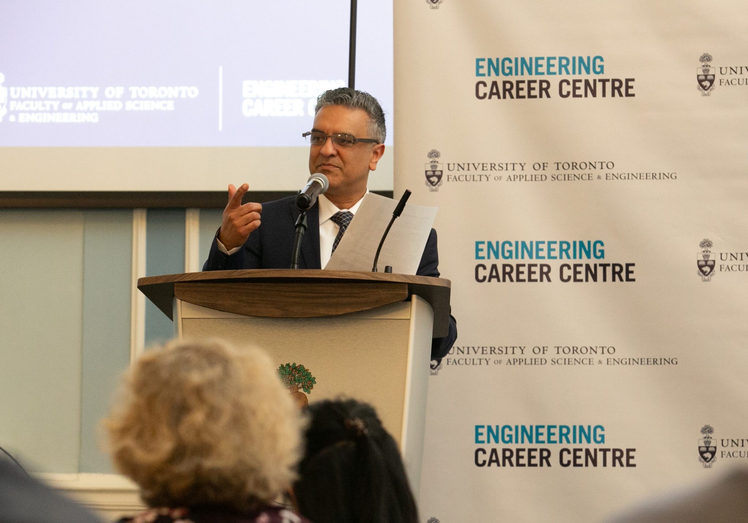 A man speaks at a podium in front of a reception crowd. Stop and repeat in background says "Engineering Career Centre."