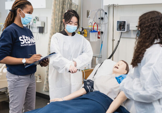 Professor Alfred and two students stand over a mannequin in a hospital bed