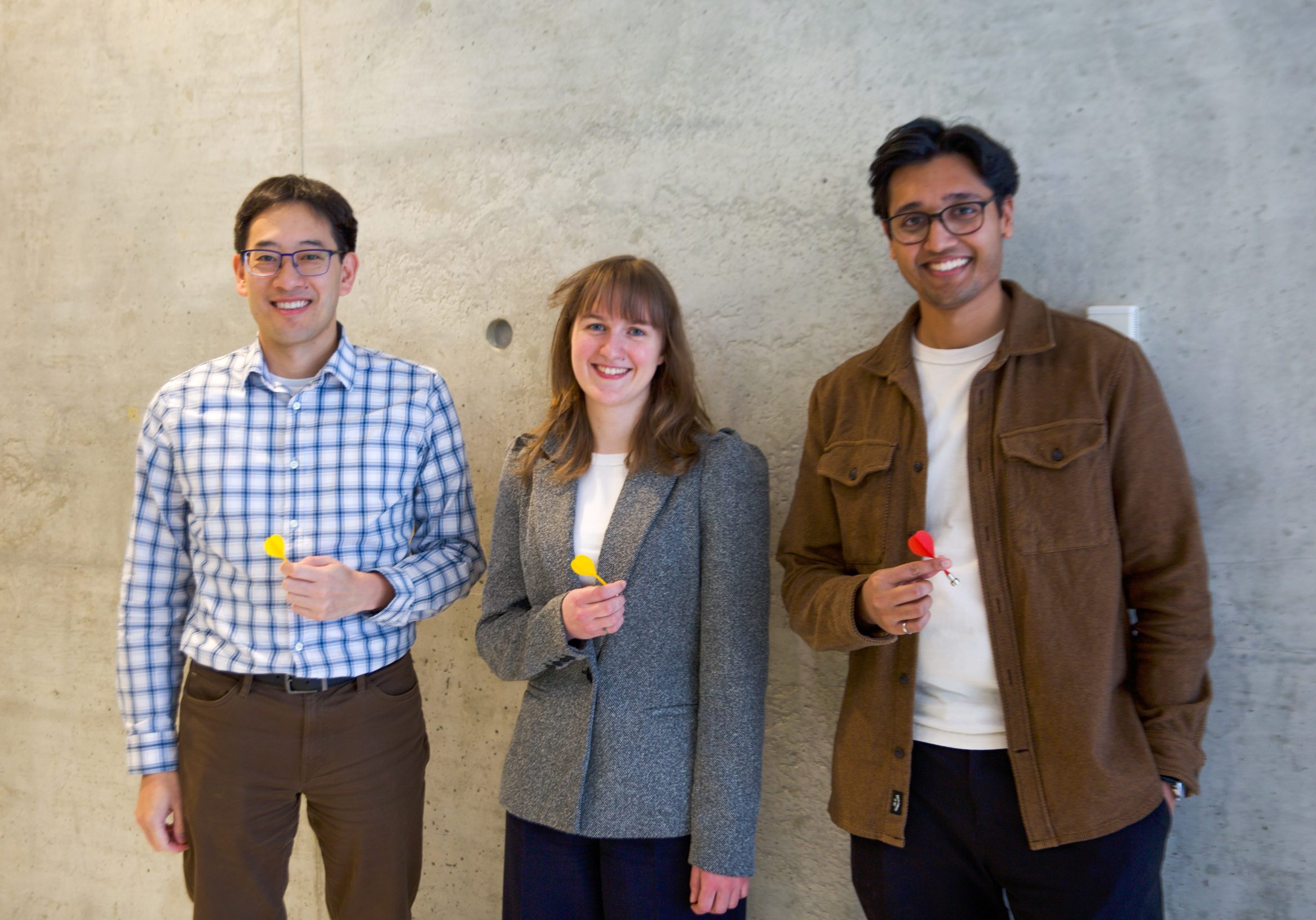 Three people stand in front of a concrete wall. Timothy Chan stands on the left wearing a shirt with a blue windowpane pattern and is holding a yellow dart, Rachael Walker stands in the centre wearing a grey blazer and is holding a yellow dart and Craig Fernandes stands on the right wearing a brown coat and is holding a red dart.