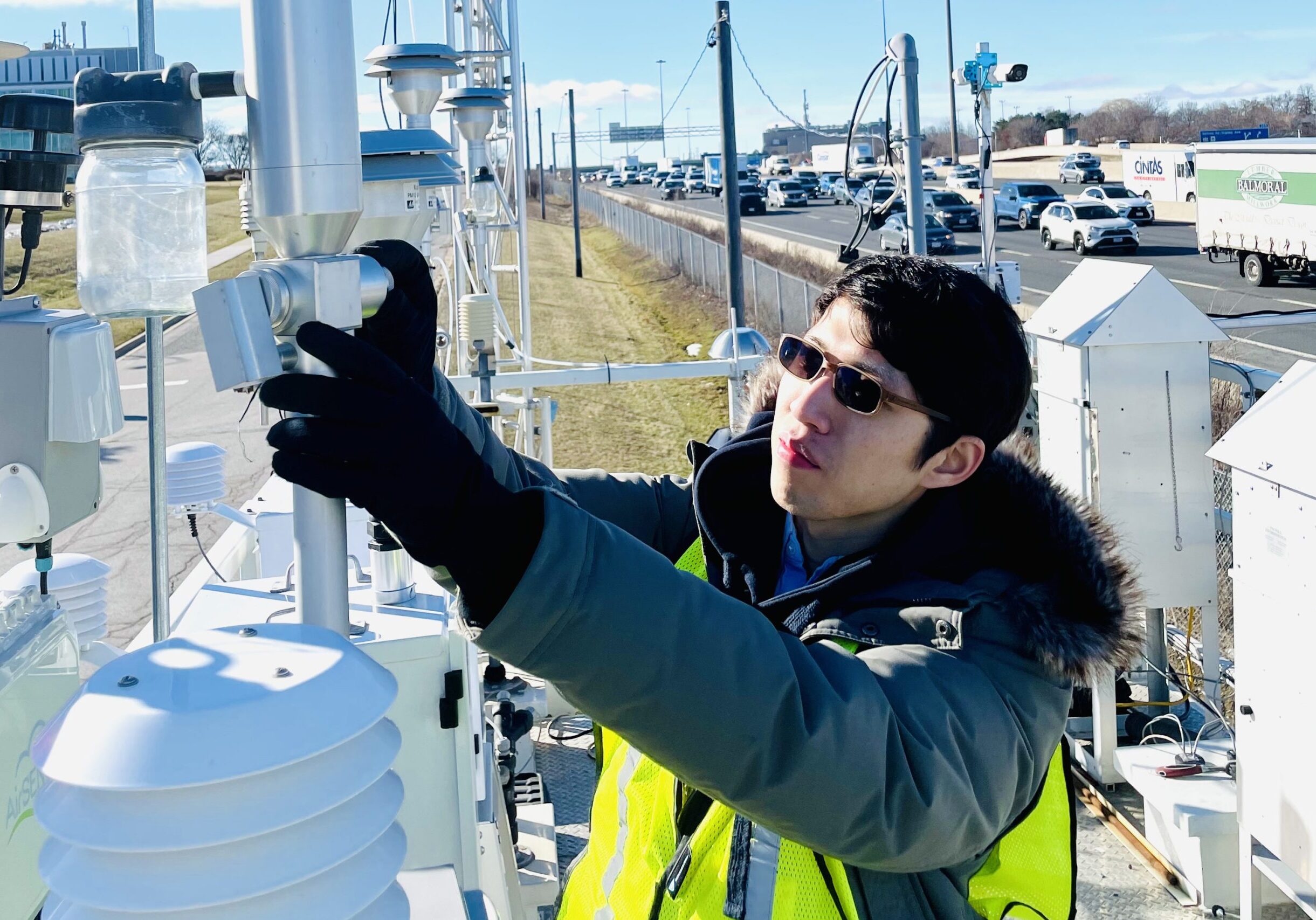 In the foreground: a researcher adjusts air quality equipment. In the background: cars are driving by, on a major roadway.
