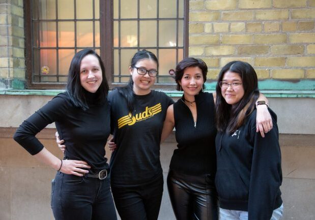 Four women stand together in front a wall with bricks and a window.